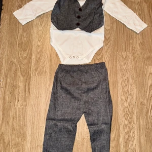 Grå kostymset med väst och rosett - Snyggt kostymset för barn med vit långärmad body och grå väst med tre bruna knappar och matchande rosett. Tillhörande grå byxor med resår i midjan. Perfekt för speciella tillfällen när man vill vara extra stilig. Cirka 2-3,5 år 