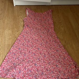 Rosa blommig klänning med öppen rygg - Supersöt rosa klänning med blommigt mönster och ärmlös design. Klänningen har en kort längd och en snygg öppen rygg med korsade band. Perfekt för sommarens alla tillfällen, lätt och skön att bära. Färgglada detaljer i blått, gult och vitt ger extra liv till looken.