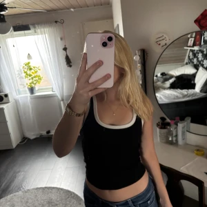 Svart ribbad linnetopp med vit kant - Säljer en svart ribbad linnetopp med breda axelband och vit kant runt halsringningen och ärmhålen. Toppen är croppad och har en tight passform som framhäver midjan. Perfekt för dig som gillar en enkel men snygg stil.