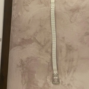 Vitt bälte med glittriga stenar - Säljer ett vitt bälte med glittriga stenar längs hela remmen och ett snyggt silverfärgat spänne med detaljer. Bältet har en rak form och är tillverkat i skinnimitation. Perfekt för att ge din outfit extra bling och stil. Några stenar saknas.
