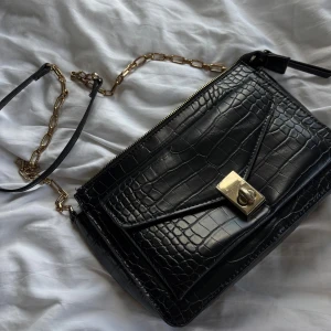 Svart axelväska, mango - Stilren svart axelväska från Mango i croco-mönstrat skinnimitation. Väskan har guldfärgade metalldetaljer, kedjerem och ett snyggt vridlås framtill. Perfekt storlek för det viktigaste och ger en lyxig känsla till din outfit. Lite rispad vid spännet, annars mycket fint skick