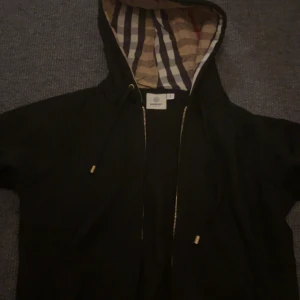 Burberry  - Svart hoodie från Burberry med klassiskt rutigt foder i huvan, guldfärgade detaljer på snörningen och dragkedja framtill. Tröjan har fickor helt ny.