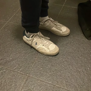 Vita sneakers från Polo Ralph Lauren. - Säljer ett par klassiska vita sneakers från Polo Ralph Lauren med blå detaljer och logga på sidan och hälen. Skorna har snörning och är tillverkade i skinn med en platt sula och rund tå. Perfekta för en clean och stilren look.