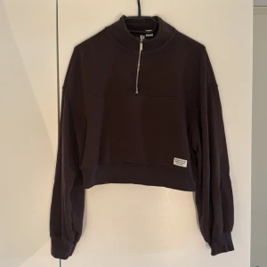Brun croppad half zip från H&M Divided - Mörkbrun croppad sweatshirt från H&M Divided med half zip-dragkedja och hög krage. Tröjan har långa ärmar och en liten patch framtill med text. Perfekt för en chill och avslappnad stil.