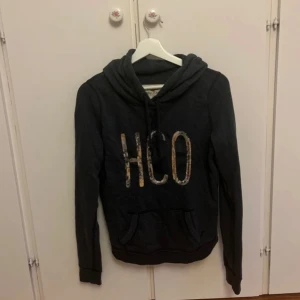 Mörkgrå hoodie från Hollister med paljetter - Säljer en mörkgrå hoodie från Hollister i storlek M. Tröjan har huva med snörning, magficka och stora HCO-bokstäver framtill i paljetter. Perfekt för dig som gillar en chill och snygg streetstil.