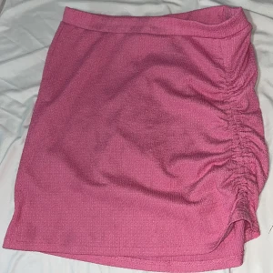 Rosa kortkjol med rynk från H&M - Säljer en rosa kortkjol som man kan  dras ut så den inte är lik kort från H&M med snygg rynkdetalj på sidan. Kjolen är gjord i ett strukturerat polyestertyg som känns mjukt och följsamt. Passformen är normal och kjolen har en enkel midjeresår för skön komfort. Perfekt för dig som gillar färg!