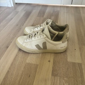 Vita Veja sneakers i skinn, strl 42 - Säljer ett par klassiska Veja sneakers i vitt skinn med beige detaljer och snörning. Skorna har den ikoniska V-loggan på sidan och Veja-tryck på hälen. Perfekta för dig som gillar stilrena och tidlösa sneakers. Storlek 42.