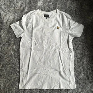 Vit t-shirt från Lyle & Scott - Vit t-shirt från Lyle & Scott med klassisk passform och korta ärmar. T-shirten har en gul broderad logga med örn på bröstet och är tillverkad i mjuk bomull. Perfekt basic-plagg med stilren look.