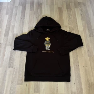 Svart hoodie Polo Bear Ralph Lauren - Svart hoodie från Polo Ralph Lauren med ikoniskt Polo Bear-tryck på bröstet. Tröjan har huva med snörning och en stor magficka. Tillverkad i mjuk bomull, perfekt för en chill och stilren look.
