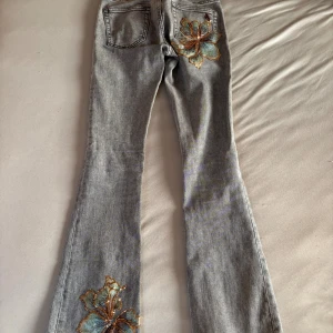 Grå bootcut jeans med paljettblommor - Grå jeans i bootcut-modell. dekorerade med stora blommor i paljetter och broderi i blått, guld och brunt på bakficka och nedre ben. Snyggt utsvängda ben och klassisk femficksdesign. Perfekta för dig som vill sticka ut med coola detaljer.