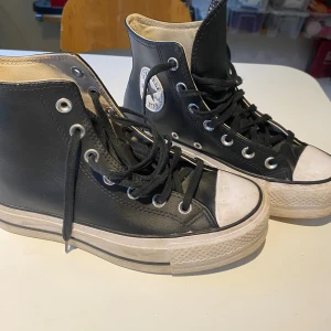 Svarta Converse Chuck Taylor All Star - Klassiska Converse Chuck Taylor All Star i svart skinn med vita detaljer och rund tå. Högt skaft, snörning med metallöljetter och vit sula. Ikonisk logga på sidan. Perfekta för en cool och avslappnad stil.