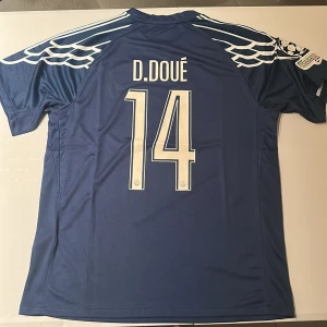 PSG blå matchtröja D. Doué #14 - Säljer en blå PSG fotbollströja med D. Doué och nummer 14 på ryggen. Tröjan har vita detaljer, vingmönster på axlarna, Qatar Airways-tryck och Jordan-logga på bröstet. Materialet är lätt och andas, perfekt för match eller träning. Replika, endast handtvätt gäller!