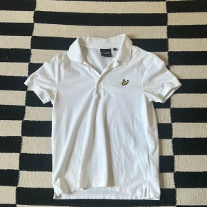 Vit Lyle & Scott pikétröja i bomull. - Fräsch vit pikétröja från Lyle & Scott med klassisk krage och korta ärmar. Tröjan har den ikoniska gula fågeln broderad på bröstet och är gjord i mjuk bomull. Perfekt för dig som gillar stilrena och enkla plagg med snygga detaljer.
