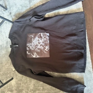 Svart sweatshirt med konsttryck H&M - Svart sweatshirt från H&M i storlek L med ett stort konstinspirerat tryck i gråskala på bröstet. Tröjan har rund halsringning och långa ärmar. Tillverkad i mjuk bomull och polyester för en skön känsla.