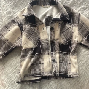 Rutig overshirt från KappAhl - Snygg rutig overshirt från KappAhl i svart, grått och beige. Jackan har klassisk krage, två bröstfickor med lock och knäppning med svarta knappar framtill. Materialet är mjukt och känns fleece-liknande, perfekt för lager på lager. Passar dig som gillar chill och trendig stil. Namnet kommer att strykas över innan de skickas iväg.