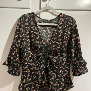 Blommig blus med volang från Bik Bok - Supersöt blommig blus från Bik Bok i svart med rosa och gröna detaljer. Djup v-ringning, knäppning framtill och volang vid ärmslut. Trekvartsärm och lätt, flowy material som känns luftigt och skönt. Plagget levereras tvättat. Finns katt i hemmet🐈