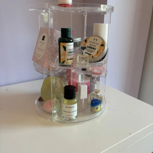 Genomskinlig sminkförvaring - Säljer en rund, roterbar sminkförvaring genomskinligmed med flera fack och hyllor. Perfekt för att organisera sminket. Enkel att montera och passar snyggt på sminkbordet.