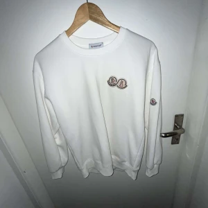 Vit tröja från Moncler - Vit sweatshirt från Moncler med broderade logopatchar på bröstet och ärmen. Tröjan har rund hals, ribbade muddar och är tillverkad i mjuk bomull. Enkel och clean design som passar perfekt till jeans eller joggers.