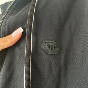 Svart hoodiejacka Emporio Armani - Svart hoodiejacka från Emporio Armani med dragkedja och luva. Jackan har ett stilrent svart logomärke på bröstet och praktiska fickor. Materialet är mjukt och känns skönt mot huden, perfekt för chill dagar. Dock har koftan en defekt som ni ser på sista bilden. 