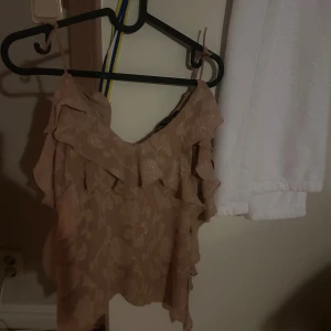 Beige volangtop från Zara - Supersöt beige linnetopp från Zara med tunna axelband och volangdetaljer framtill. Toppen har ett subtilt blommönster i tyget och är tillverkad i ett lätt, glansigt material som känns lyxigt. Perfekt för dig som gillar romantiska och feminina plagg. Förra älgen klippte sönder axelbanden tyvärr men kan använda utmärkt ändå! 