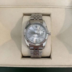 Datejust silverfärgad klocka - Säljer en stilren Datejust med silverfärgad urtavla och klassisk räfflad bezel. Klockan har en länk i rostfritt stål och datumfönster vid klockan tre. Levereras i originalbox med tillbehör. Perfekt för dig som gillar exklusiva accessoarer.