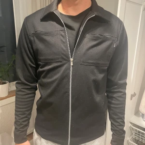 Svart overshirt med dragkedja från Jack and Jones - Stilren svart overshirt med hel dragkedja från Jack and Jones. Lite knopprig men annars bra skick.