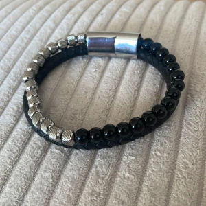 Svart och silver armband från Vezzi  - Stilrent armband från Vezzi med svart flätat band, svarta glansiga pärlor och detaljer i silverfärgad metall. Magnetlås med Mezz-logga. Perfekt accessoar för en modern look.