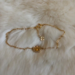 Guldigt armband från Tory Burch - Snyggt och stilrent armband från Tory Burch i guldtonad metall. Armbandet har en tunn kedja och ett runt hänge med den ikoniska Tory Burch-loggan, omgiven av små glittrande stenar. Perfekt accessoar för att lyfta din outfit.