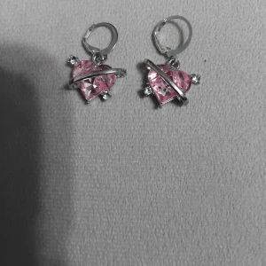 Rosa hjärtformade örhängen med stenar - Supersöta örhängen med hjärtformade rosa stenar och silverfärgade detaljer. Hängena har en glittrig känsla med små klara stenar och en unik design där en silverfärgad båge går över hjärtat. Perfekta för att ge din look en söt och trendig touch.