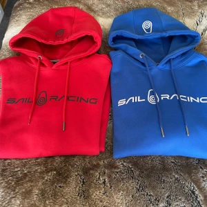 Sail Racing hoodie i rött och blått - Två snygga hoodies från Sail Racing, en i rött och en i blått. Båda har huva med snörning, tryckt logga framtill och mjukt material. Perfekt för dig som gillar sportig och avslappnad stil. Finns en defekt på snöret på den röda. Paketpris 900kr/ 500krst/ 450kr