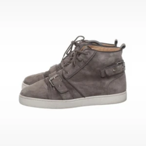  Christian Louboutin - Snygga gråa sneakers från Christian Louboutin i mocka med vita sulor och klassisk röd undersida. Skorna har snörning och coola spänndetaljer på sidorna för en edgy vibe. Perfekta för dig som vill sticka ut med exklusiva och trendiga skor.
