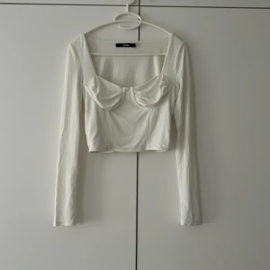 Vit långärmad korsettopp Bik Bok - Säljer en vit långärmad korsettopp från Bik Bok i storlek S. Toppen är croppad med markerade kupor och fyrkantig ringning. Tillverkad i mjukt stretchigt material som sitter snyggt på kroppen. Perfekt att styla med jeans eller kjol för en trendig look.