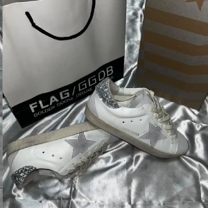 Golden Goose Superstar sneakers med glitter - Golden Goose Superstar sneakers i vitt skinn med grå mockadetaljer och en stor grå stjärna på sidan. Hälpartiet är täckt av silverglittrande material som ger extra edge. Skorna har snörning och platt sula, perfekt för dig som gillar att sticka ut med din stil.