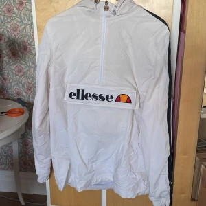 Vit vindjacka från Ellesse med logga - Vit vindjacka från Ellesse med stor logga framtill och halv dragkedja. Jackan har huva, svarta detaljer längs ärmarna och meshfoder på insidan. Perfekt för en sportig och avslappnad stil.