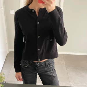 Svart kofta med knappar och fickor - Säljer en svart, klassisk kofta med rund halsringning och knäppning framtill. Koftan har två fickor framtill och är långärmad. Den är i ett mjukt, stickat material och har en rak, något croppad passform. Perfekt att slänga över en t-shirt eller linne.