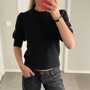 Svart stickad topp med puffärm - Säljer en svart stickad topp med rund hals och korta puffärmar. Modellen har ribbade kanter vid hals och nederkant. Perfekt för dig som gillar en enkel men trendig look.