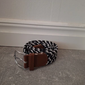 Svartvitt flätat bälte med skinn - Snyggt flätat bälte i svart och vitt med bruna skinn-detaljer och silvrigt spänne. Bältet har ett randigt mönster och passar perfekt till jeans eller chinos för en stilren look. Flexibelt material som gör det enkelt att justera längden.