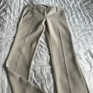 Beiga kostymbyxor från BikBok - Snygga beiga kostymbyxor från BikBok i storlek 34. Byxorna har hög midja, raka ben och pressveck framtill. De är tillverkade i ett mjukt material med skön passform och har hällor för bälte samt fickor både fram och bak.