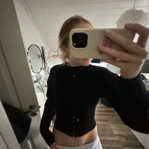 Svart långärmad korsett-topp - Säljer en svart långärmad korsett-topp med snygga metallhakar framtill. Toppen är tight och har en croppad passform som framhäver midjan. Perfekt för dig som vill ha en edgy och trendig look.