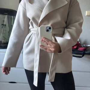 Beige omlottkappa med bälte - Snygg beige kappa med bred krage och knytbälte i midjan. Jackan har en omlottmodell och långa ärmar, perfekt för dig som gillar stilrena och enkla plagg. Materialet känns mjukt och har en lyxig känsla. Passformen är normal och ger en clean look.