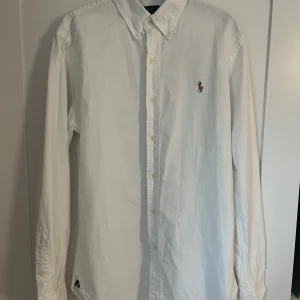 Vit skjorta från Polo Ralph Lauren - Klassisk vit skjorta från Polo Ralph Lauren i slim fit-modell. Skjortan har lång ärm, knappar hela vägen och broderad logga på bröstet. Tillverkad i bomull och har en stilren, tidlös look med button down-krage.