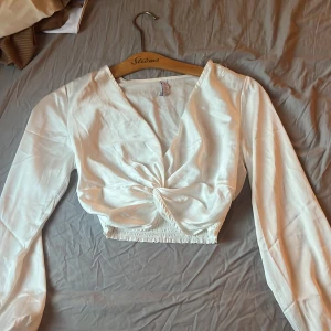 Vit blus med lång ärm - Vit tunn blus från Amisu med croppad passform och lång ärm. Blusen har snygg twistad detalj framtill och resår vid midjan. Materialet är glansigt och känns lyxigt, perfekt för att lyfta din outfit.