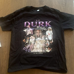 Svart T-Shirt - Lil Durk - Svart t-shirt med stort  tryck av Lil Durk och texten 'The Voice' på ryggen. Framsidan har motiv med flera bilder, lila och vita detaljer samt fjärilar. Klassisk passform med rund hals och korta ärmar. 