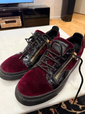 Vinröda Giuseppe Zanotti - Säljer ett par lyxiga vinröda sneakers från Giuseppe Zanotti med svart lackdetalj, gulddragkedjor på sidorna och svarta snören. Skorna har platt sula och är tillverkade i sammet och skinn, vilket ger en riktigt exklusiv vibe. Perfekt för dig som gillar att sticka ut med din stil.