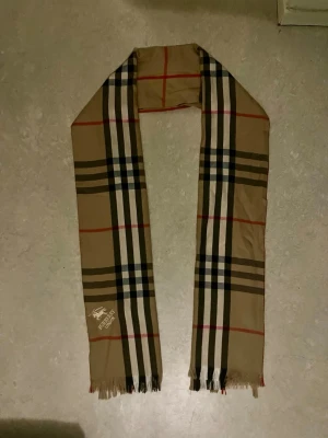 Vintage Burberry halsduk - Säljer en klassisk vintage beige halsduk från Burberry med det ikoniska rutiga mönstret i svart, vitt och rött. Halsduken har fransar i kanterna och Burberry London-logga broderad i vitt. Perfekt accessoar för att lyfta din outfit.