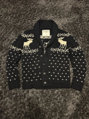 Abercrombie & Fitch Cardigan - Säljer en svart stickad kofta från Abercrombie & Fitch i storlek M. Koftan har krage, knappar framtill och ett snyggt vintermönster med vita älgar och små detaljer. Materialet är mestadels bomull med inslag av ull och polyester. Perfekt för kyliga dagar med ett juligt mönster!                         Hör gärna av dig vid frågor!