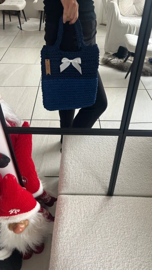 Mörkblå stickad handväska med rosett passar till laptop!  - Unik handväska i mörkblå och blå stickad design, med två rejäla handtag och en vit rosett framtill som detalj. Väskan är rektangulär och har ett öppet fack utan dragkedja. Perfekt för dig som gillar handgjorda accessoarer med personlig touch.