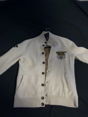 Biege kofta från Morris med brodyr - Säljer en beige kofta från Morris i storlek S med snygg broderad logga och text på ryggen och ärmen. Koftan har svarta knappar, ribbade muddar och två fickor framtill. Perfekt för dig som gillar klassisk stil med detaljer.