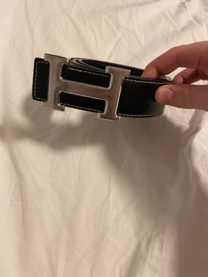 Svart skärp med H-spänne i skinn - Säljer ett svart skärp i äkta skinn med ett stort, silverfärgat H-spänne. Skärpet har vit kontrastsöm längs kanterna och är bredare än vanliga bälten, vilket ger en riktigt statement look. Perfekt accessoar för att lyfta vilken outfit som helst.