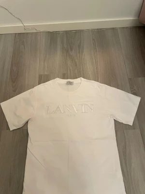 Vit Lanvin Paris t-shirt i bomull - Säljer en clean vit t-shirt från Lanvin Paris i storlek M. T-shirten har korta ärmar och ett broderat Lanvin Paris-logo framtill. Materialet är 100% bomull och den har en klassisk rund hals. Perfekt för dig som gillar stilrena designer och exklusiva märken.